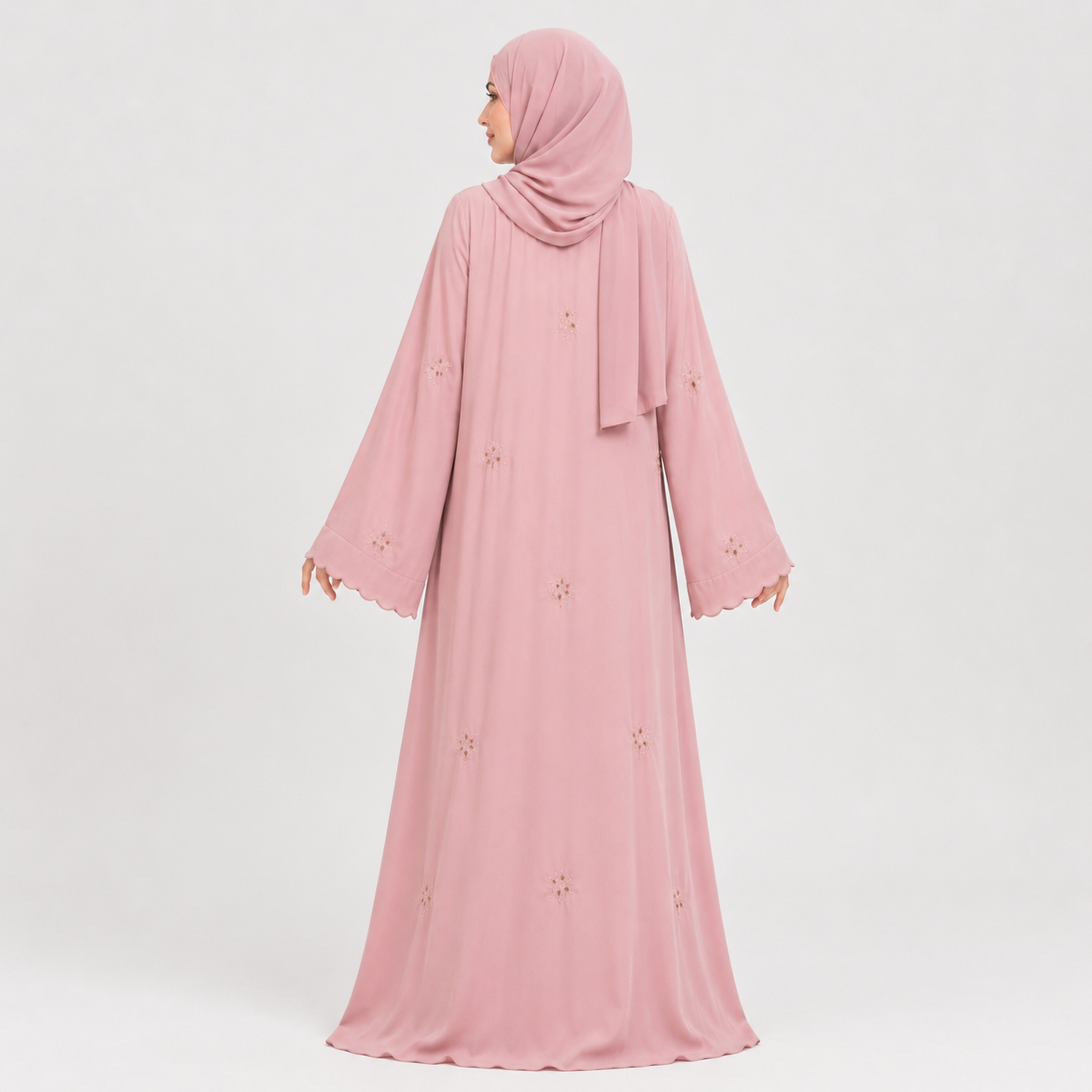 A woman wearing a long pink abaya dress with a matching hijab, shown from the back against a light gray studio background. The dress has long sleeves, a modest silhouette, and small embroidered floral details scattered across the fabric.
