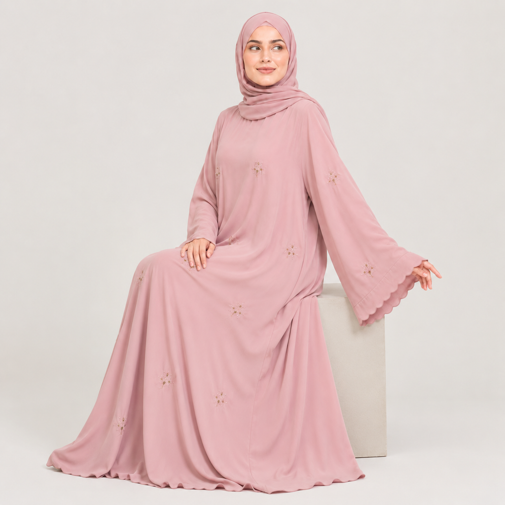 A woman wearing a long pink abaya dress with a matching hijab, shown from the back against a light gray studio background. The dress has long sleeves, a modest silhouette, and small embroidered floral details scattered across the fabric.