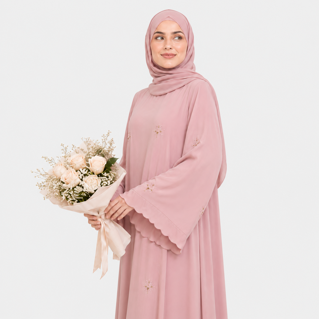 A woman wearing a long pink abaya dress with a matching hijab, shown from the back against a light gray studio background. The dress has long sleeves, a modest silhouette, and small embroidered floral details scattered across the fabric.