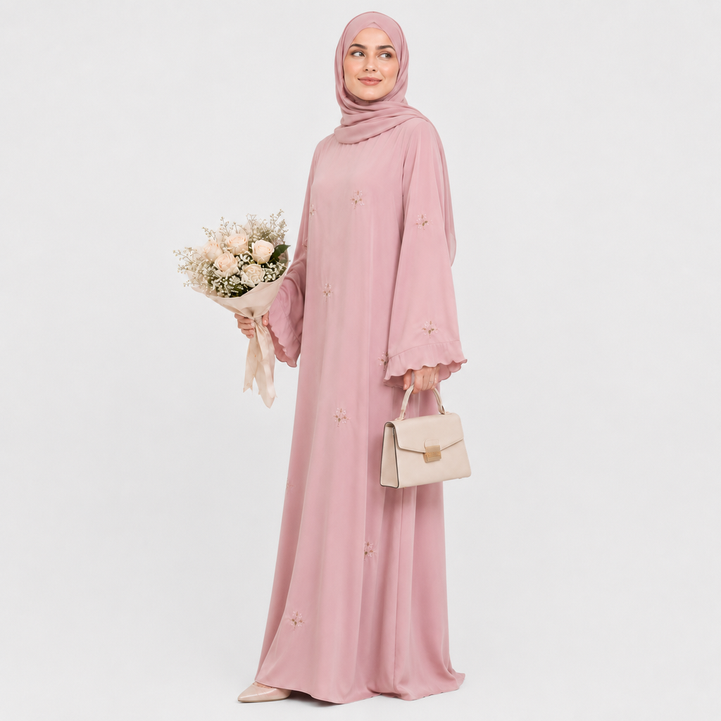 A woman wearing a long pink abaya dress with a matching hijab, shown from the back against a light gray studio background. The dress has long sleeves, a modest silhouette, and small embroidered floral details scattered across the fabric.