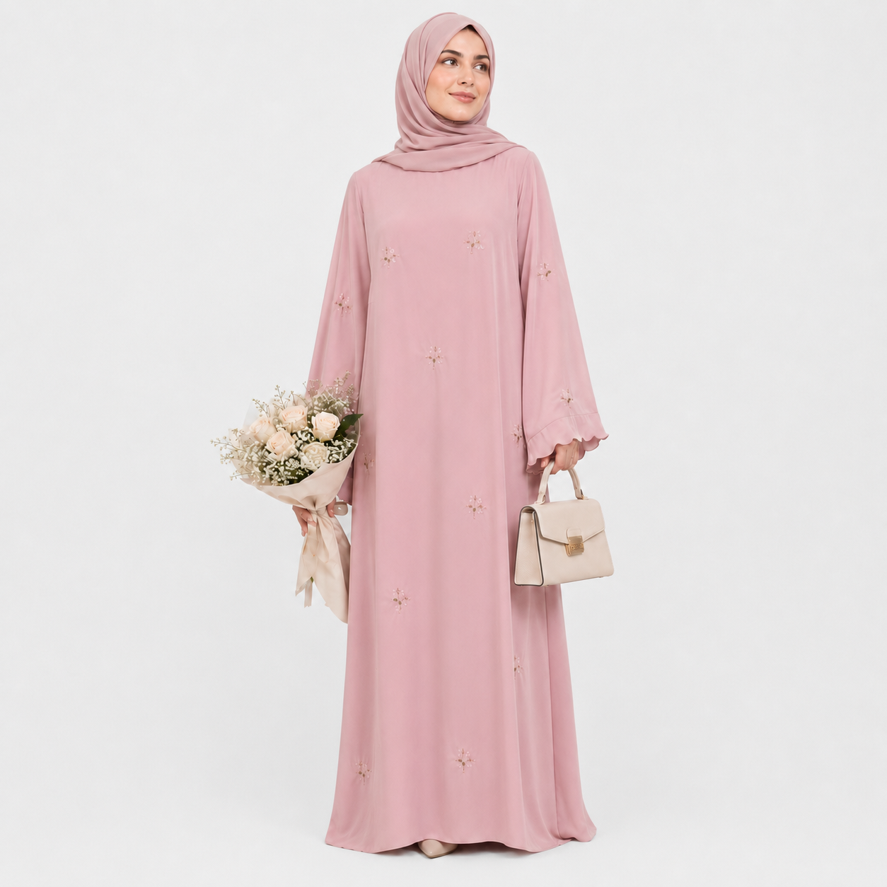 A woman wearing a long pink abaya dress with a matching hijab, shown from the back against a light gray studio background. The dress has long sleeves, a modest silhouette, and small embroidered floral details scattered across the fabric.