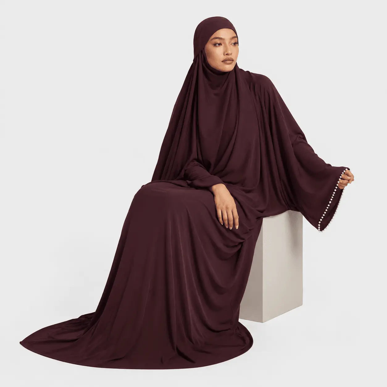 Woman wearing a burgundy abaya sitting on a white pedestal against a light gray background