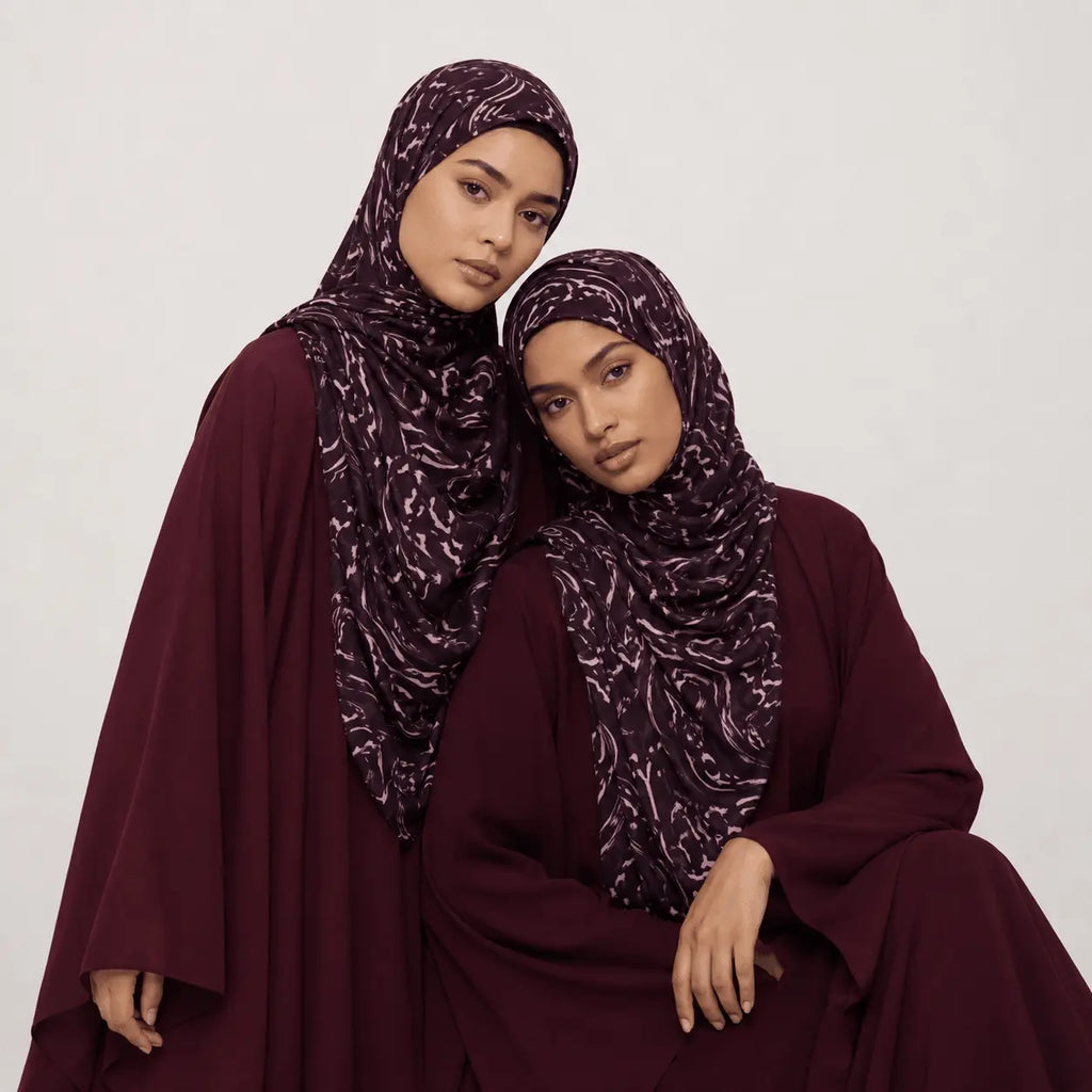 Two women wearing matching burgundy abayas with patterned hijabs on a white background
