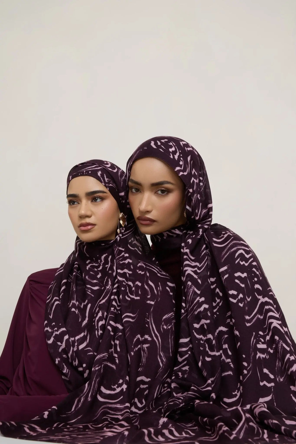 Two women wearing purple patterned headscarves against a white background