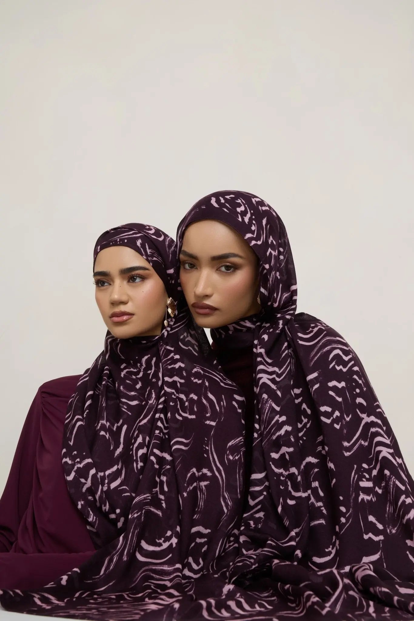 Two women wearing purple patterned headscarves against a white background