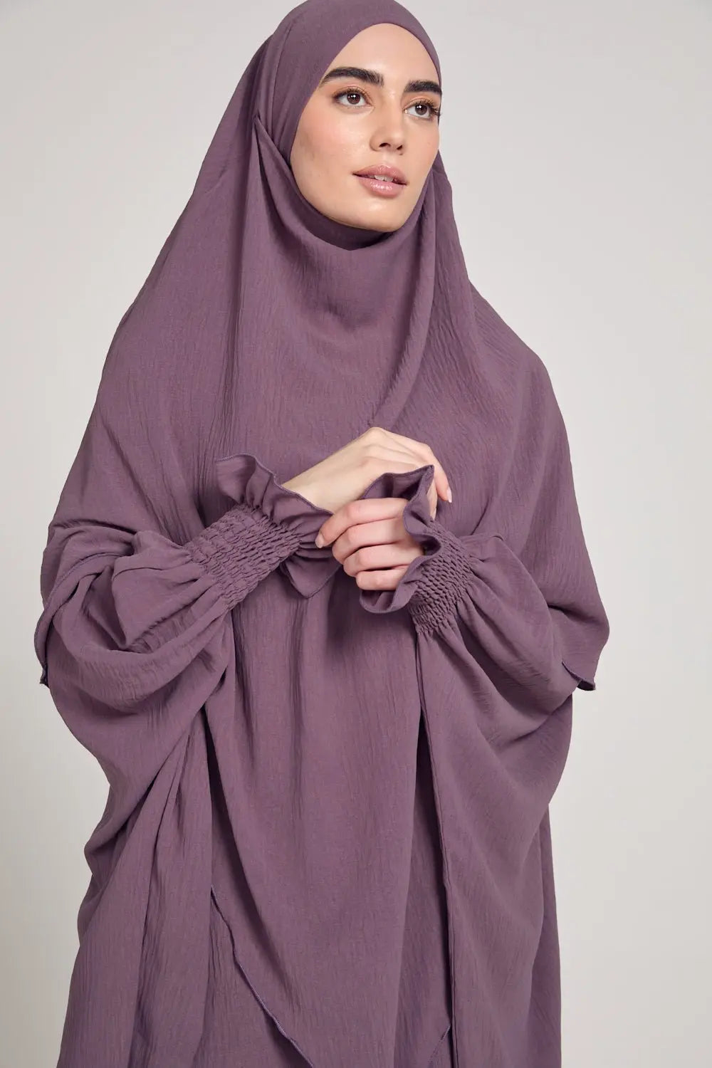 Woman wearing a purple hijab and dress against a plain background