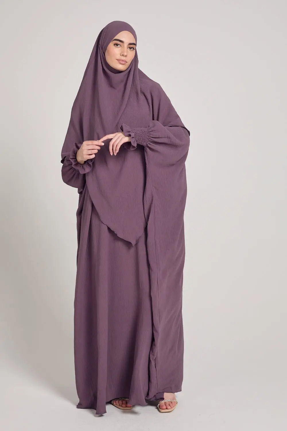 Woman wearing a long, flowing purple dress and hijab on a plain background