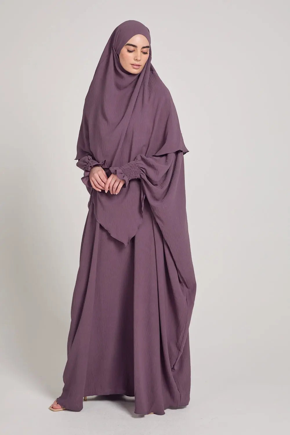 Woman wearing a long, flowing purple dress with a matching hijab on a plain background