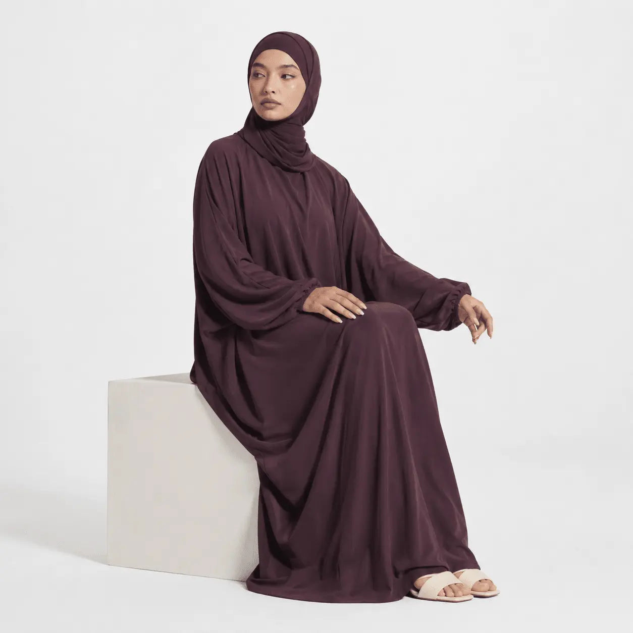 Woman wearing a long, dark purple dress sitting on a white block against a white background
