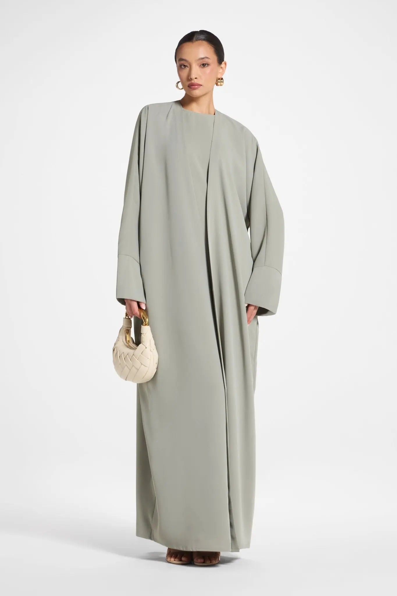 Woman wearing a long, light green dress holding a beige handbag on a white background