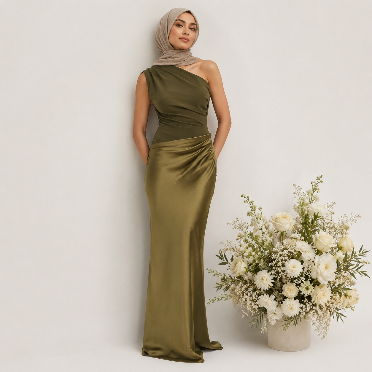 Woman in an olive green one-shoulder gown standing next to a floral arrangement on a white background