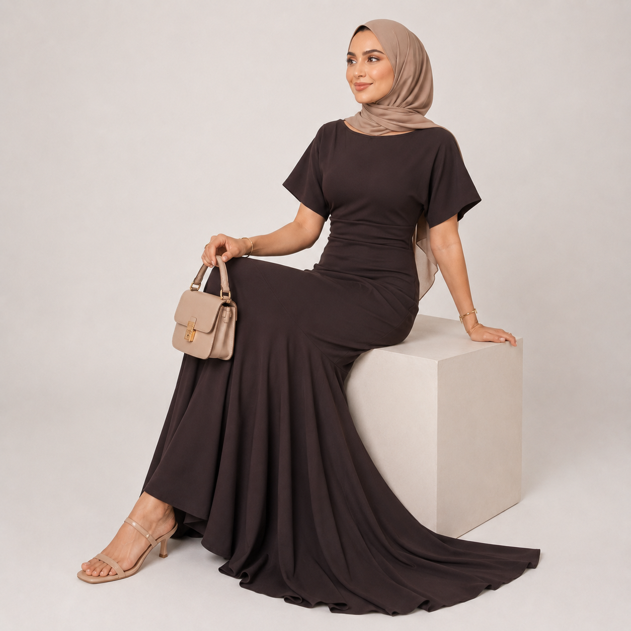 Woman in a black dress and beige hijab sitting on a white cube against a light gray background
